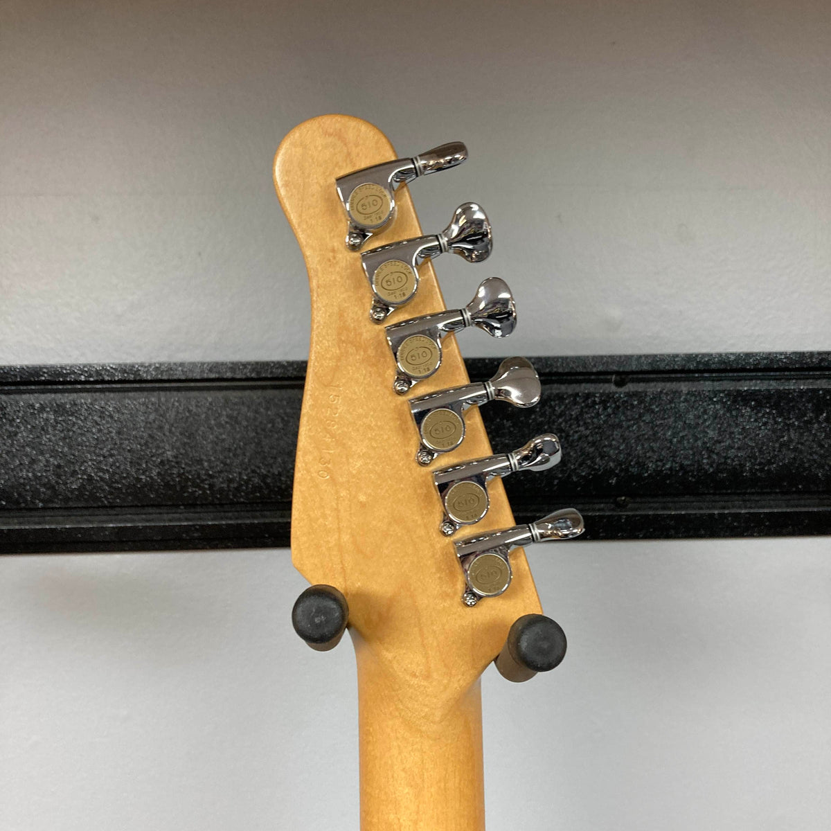 Close-up of the back of a Godin Passion Custom Swamp Ash Electric Guitar w/ GigBag Consign headstock, featuring six chrome tuning pegs, mounted on a wall hanger against a plain background.