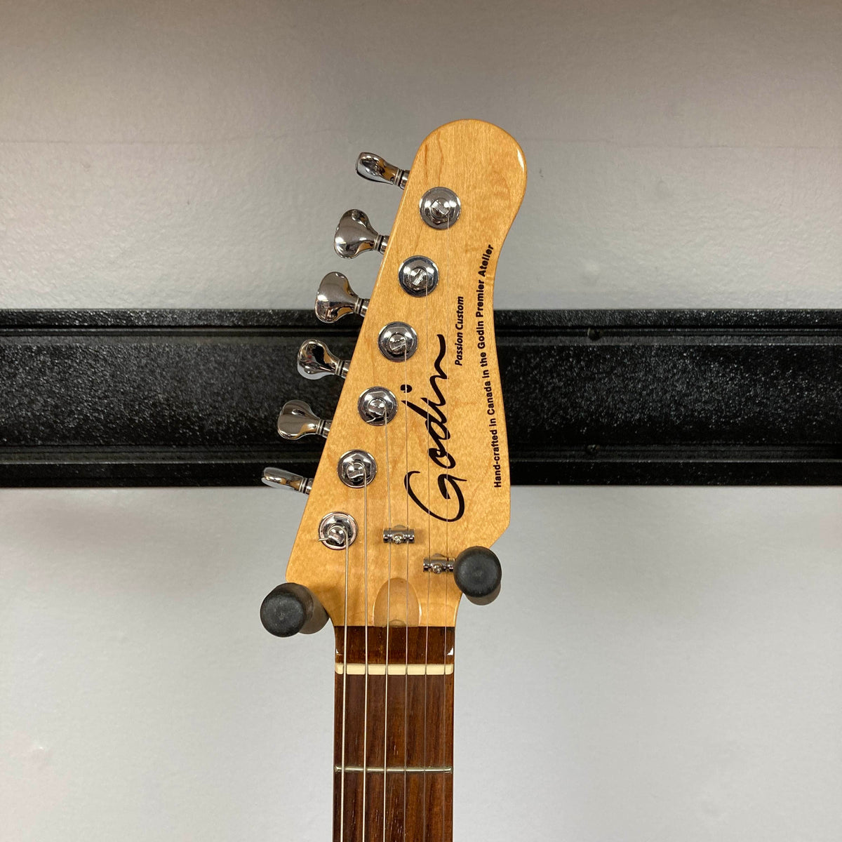 Headstock of a Godin Passion Custom Swamp Ash Electric Guitar w/ GigBag Consign by Godin, with tuning pegs visible, mounted on a wall rack against a plain white background. Renowned for its remarkable tonal versatility, this model often incorporates Seymour Duncan P-Rail pickups, enhancing its dynamic sound capabilities.