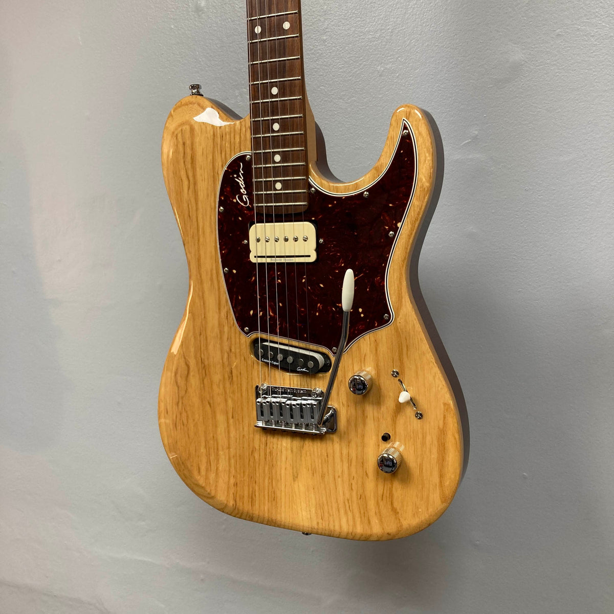 A natural-finish Godin Passion Custom Swamp Ash electric guitar with a tortoiseshell pickguard, two pickups, three control knobs, and a tremolo bridge hangs elegantly against a light gray wall—showcasing the exceptional tonal versatility of this Godin instrument.