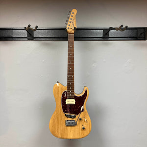 The Godin Passion Custom Swamp Ash Electric Guitar w/ GigBag Consign, by Godin, features a natural wood finish, dark red pickguard, and six tuning pegs. It hangs on a wall-mounted guitar rack against a light gray wall and is equipped with Seymour Duncan P-Rail pickups for exceptional tonal versatility.