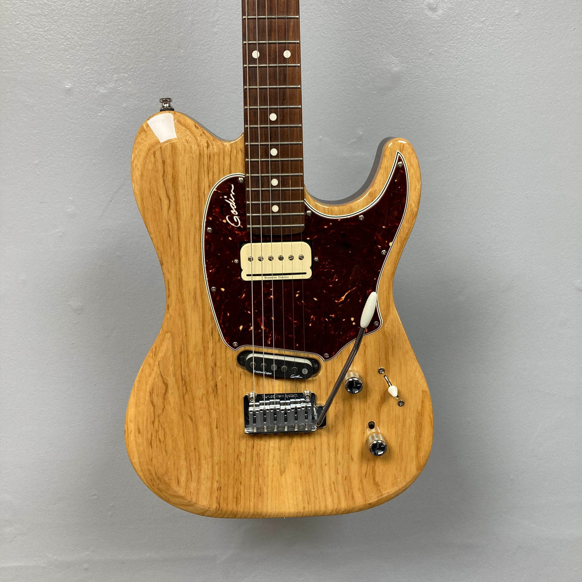 Godin Passion Custom Swamp Ash Electric Guitar w/ GigBag Consign by Godin, featuring a natural wood finish, dark red pickguard, single-coil pickup, and a whammy bar, mounted against a gray wall. Offering tonal versatility inspired by instruments like the Godin Passion Custom Swamp Ash or pickups such as Seymour Duncan P-Rail, this guitar is ideal for players seeking dynamic sound options and eye-catching aesthetics.