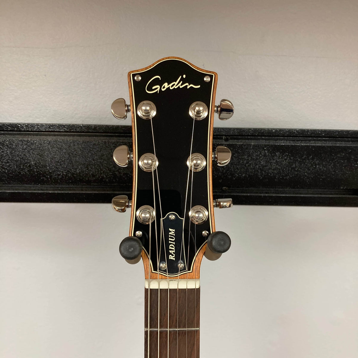 Close-up of the headstock of the Godin Radium Matte Green guitar, featuring tuning pegs, the Godin logo, and part of the neck against a plain wall background.