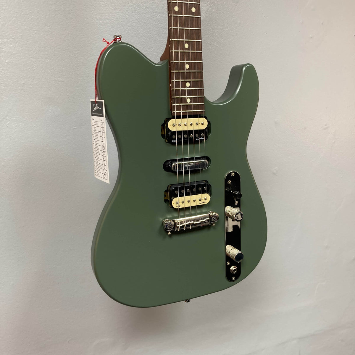 A Godin Radium Matte Green electric guitar with Godin Zebra Humbuckers (H-S-H pickup configuration), tag on the headstock, hangs against a light gray wall.