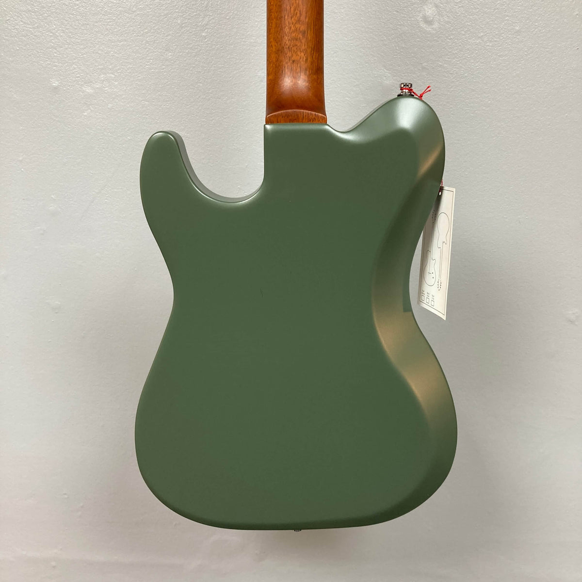 Back view of a Godin Radium Matte Green electric guitar with a brown neck and Godin Zebra Humbuckers, hanging on a plain white wall; a white tag is attached near the neck joint.