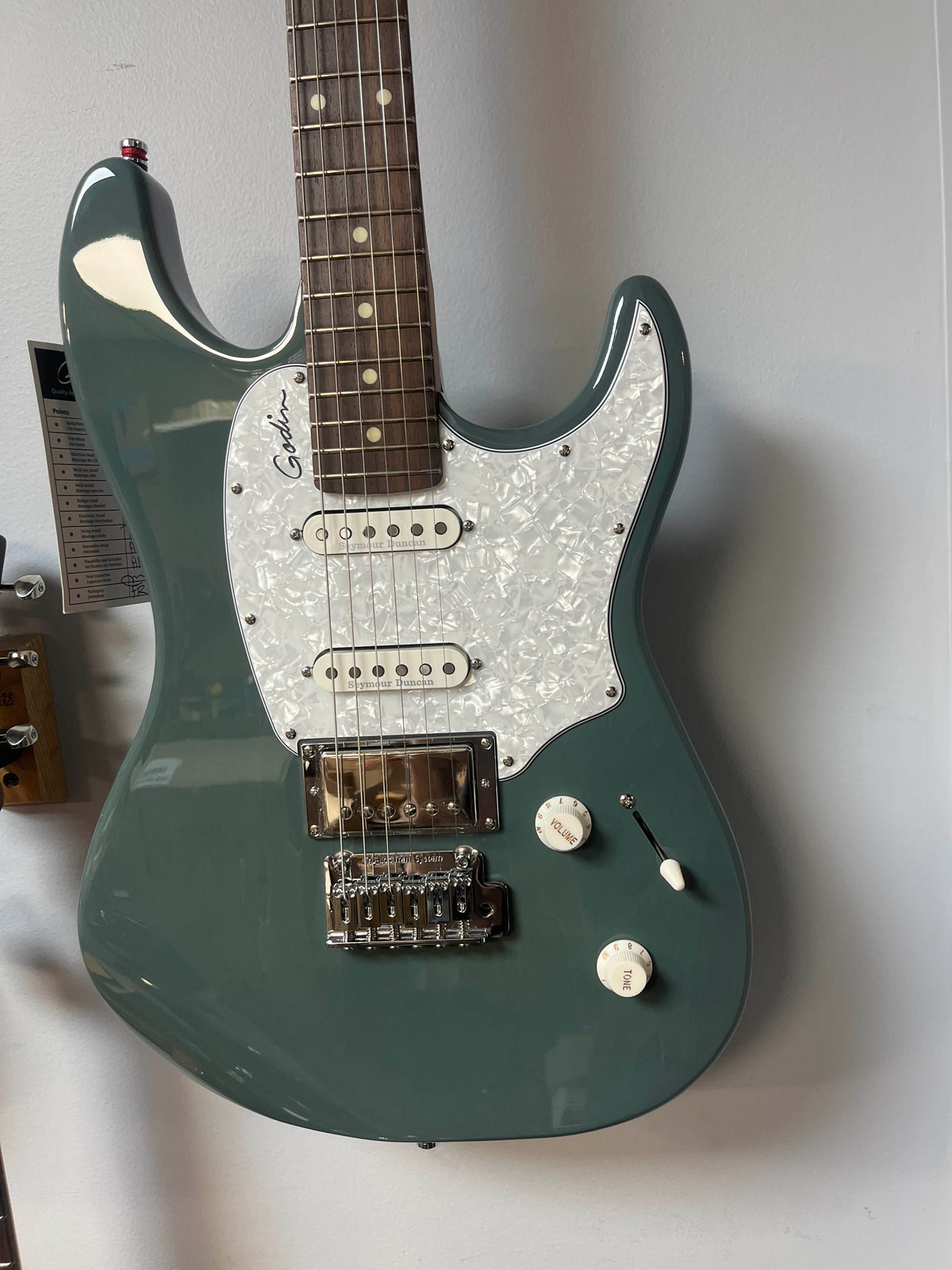 A green Godin Session T-Pro with Bag electric guitar by Godin, featuring an H-S-S pickup configuration, Seymour Duncan pickups, white pearloid pickguard, two control knobs, and a three-way switch hangs on the wall.