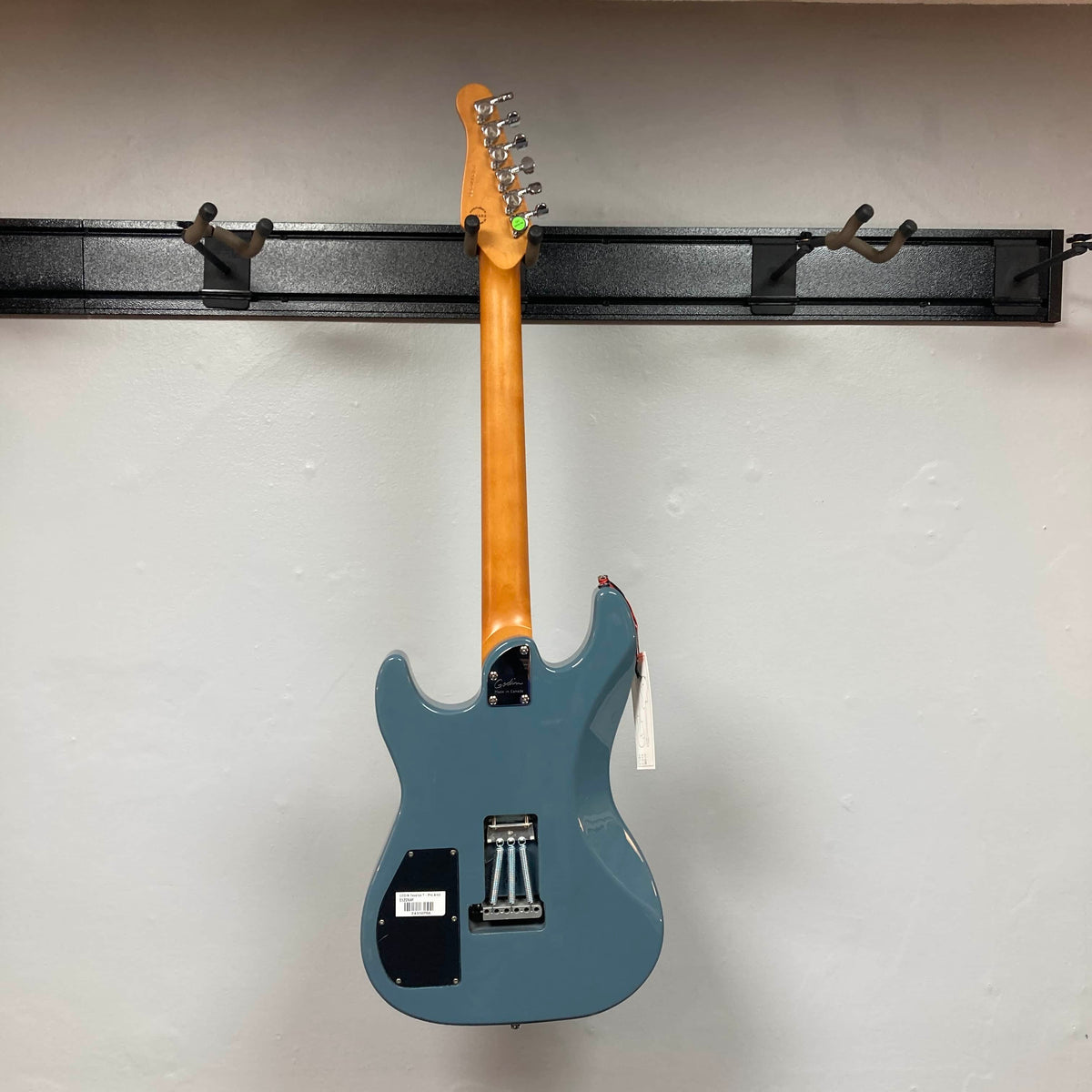 A Godin Session T-Pro with Bag electric guitar (blue, H-S-S pickups) by Godin hangs from a black wall-mounted rack, shown from the back against a plain white wall.