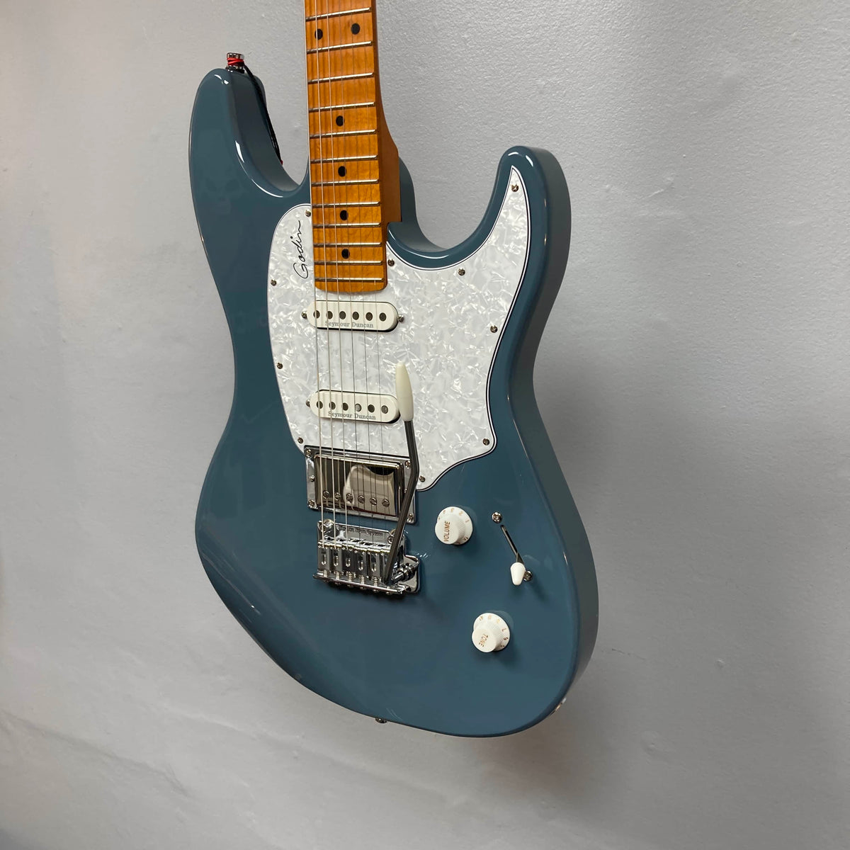 A Godin Session T-Pro electric guitar with a blue finish, white pickguard, H-S-S pickups, three knobs, and a maple neck from Godin is mounted on a plain light wall.
