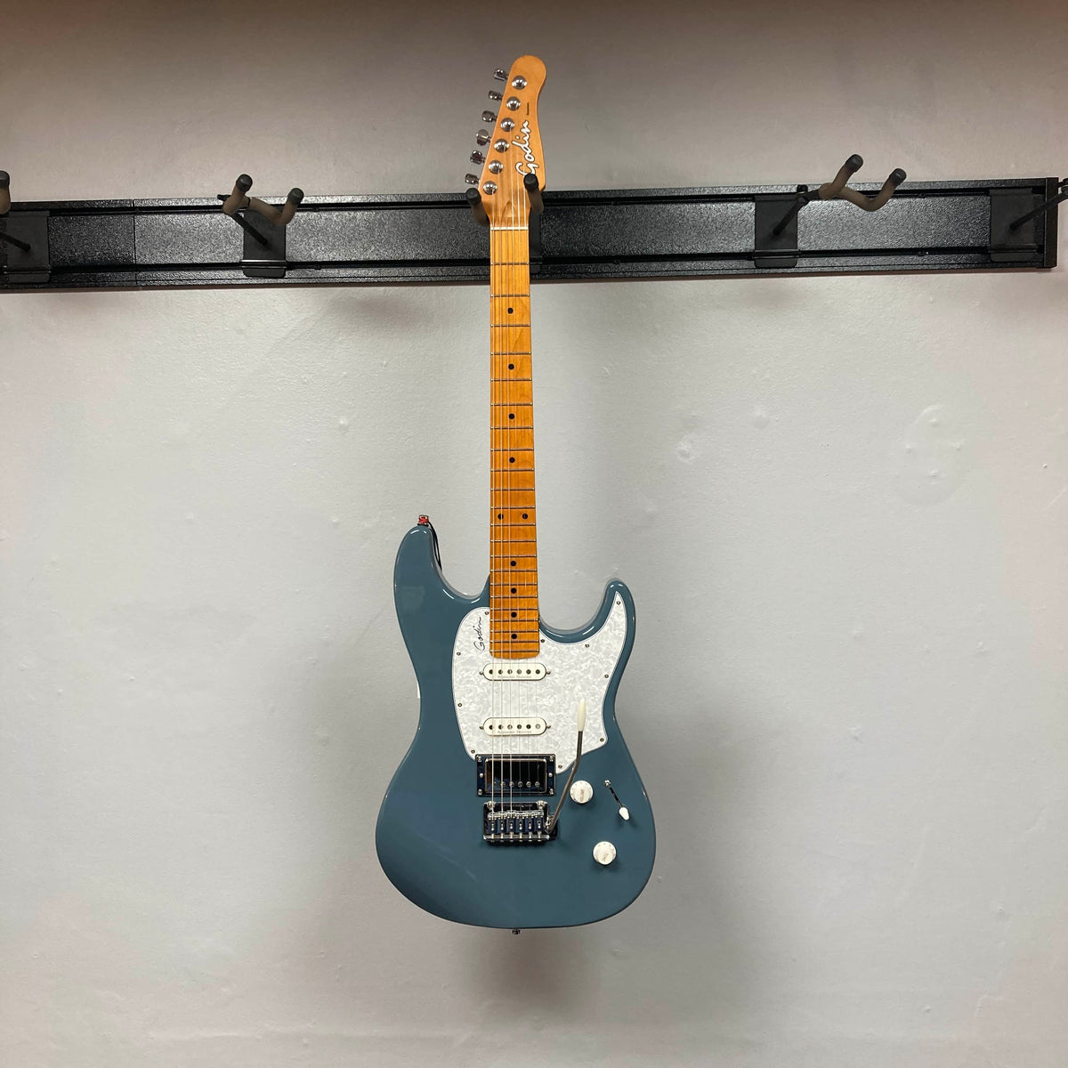 A Godin Session T-Pro with Bag electric guitar by Godin, featuring a blue finish, maple neck, white pickguard, and H-S-S pickups, hangs on a black wall-mounted rack against a plain light-colored wall.