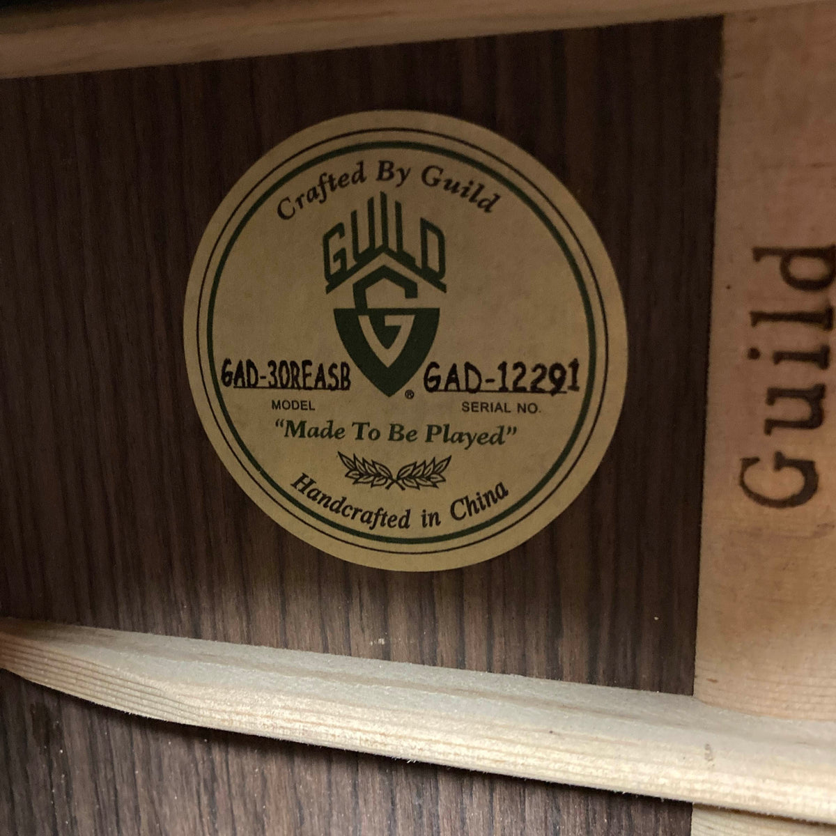 A close-up of a Guild Gad-30REASB w/Case guitar label inside the body displays the model GAD-30REASB, serial number GAD-12291, and the phrase "Made To Be Played," indicating this orchestra-sized acoustic-electric guitar by Guild was handcrafted in China with solid Indian rosewood back and sides, and is equipped with a premium Fishman Matrix pickup.