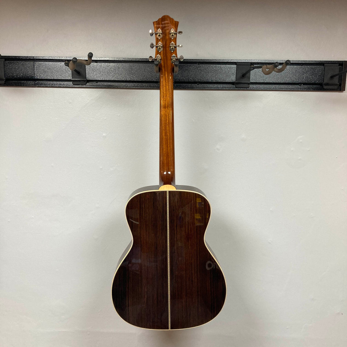 The Guild Gad-30reasb w/Case acoustic-electric guitar by Guild is displayed hanging on a wall, viewed from the back to showcase its dark solid Indian rosewood finish and metal tuning pegs.
