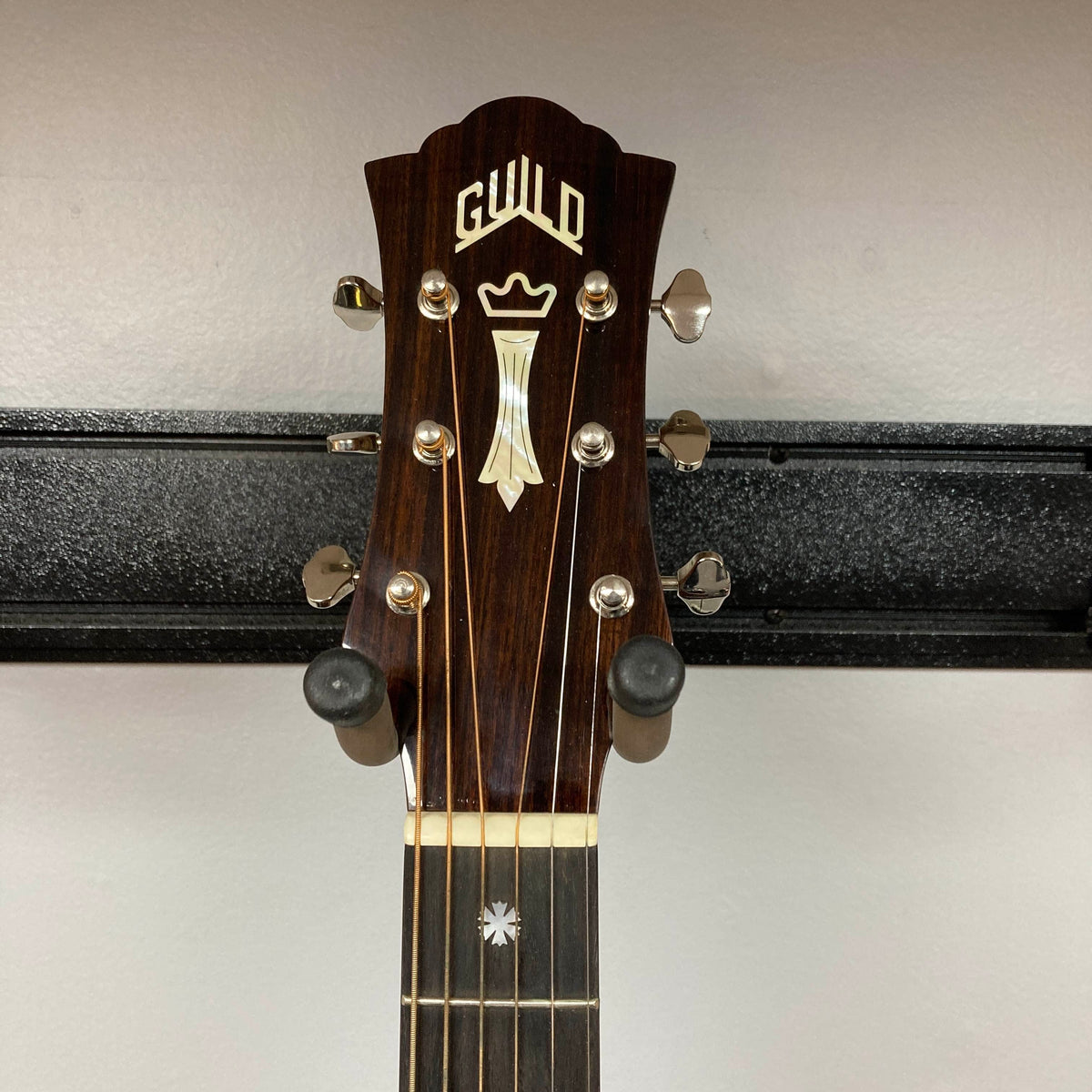 Headstock of a Guild Gad-30reasb acoustic-electric guitar, crafted from solid Indian rosewood, featuring tuning pegs and a decorative inlay, mounted on a wall hanger.