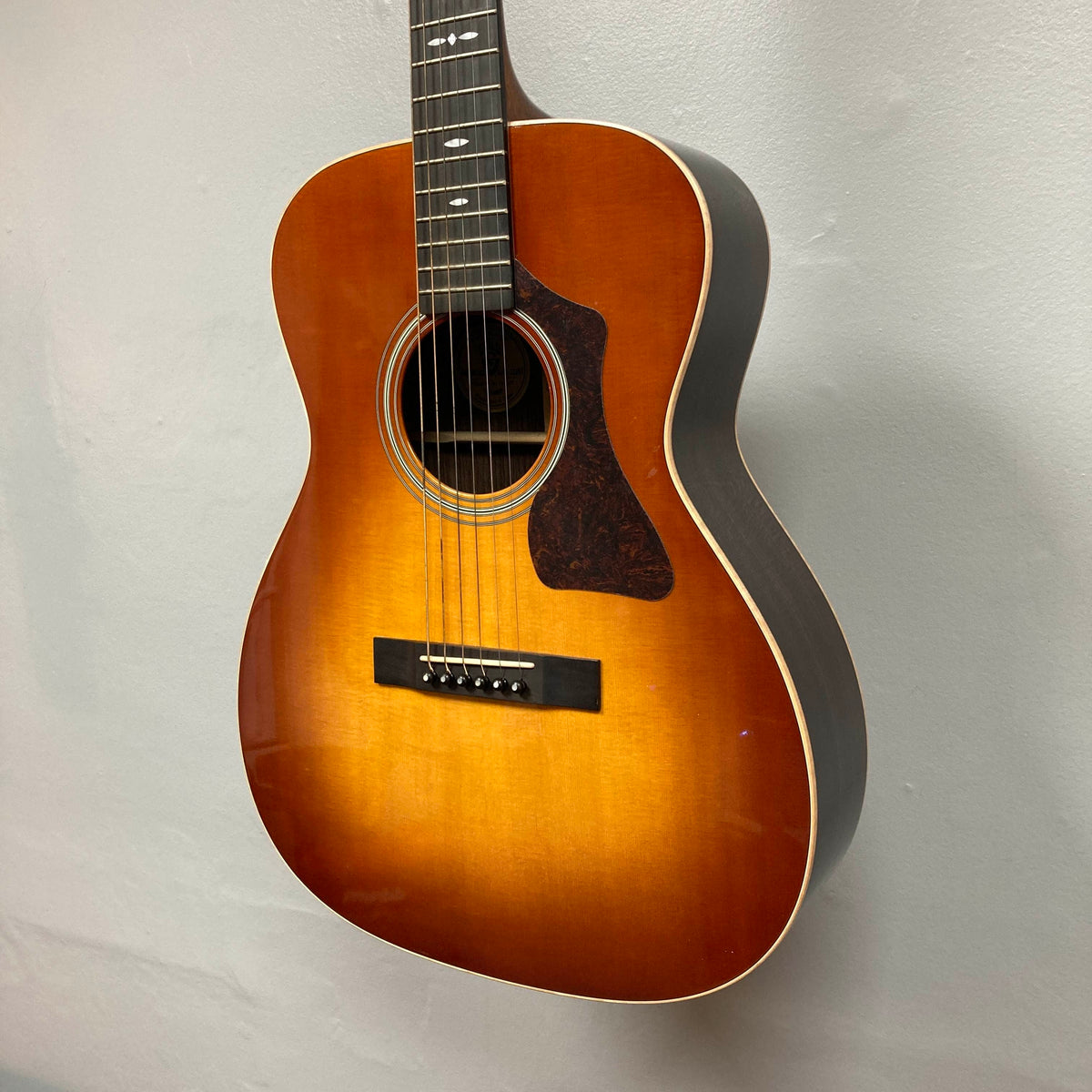 A Guild Gad-30reasb w/Case acoustic-electric guitar by Guild, featuring a classic sunburst finish, hangs on a light-colored wall. This orchestra-size model offers solid Indian rosewood back and sides, six strings, a tortoiseshell pickguard, and comes equipped with a Fishman Matrix pickup for exceptional amplified sound.