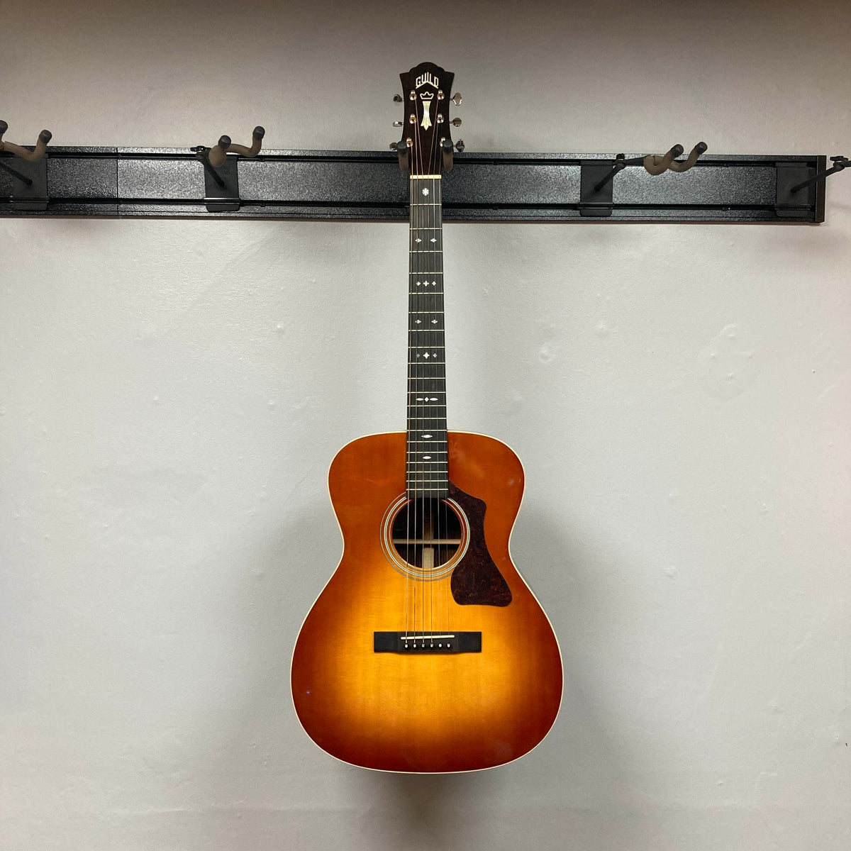 A Guild Gad-30reasb w/Case, an orchestra-size acoustic-electric guitar with a striking sunburst finish, hangs on a black wall-mounted rack against a plain white wall. Its solid Indian rosewood back and sides catch the light, while the discreet Fishman Matrix pickup ensures this Guild instrument is both visually stunning and performance-ready.