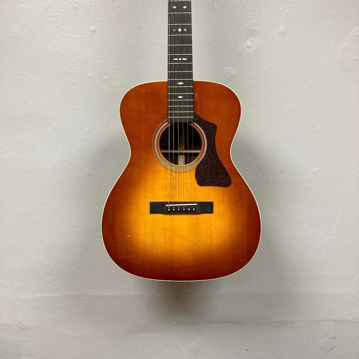 The Guild Gad-30reasb w/Case, an orchestra-size acoustic-electric guitar with a sunburst finish, is hanging on a plain, light-colored wall and features a Fishman Matrix pickup for exceptional amplified sound.