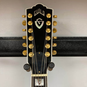 Close-up of a black Guild USA F-512 w/OHSC 2005 Circa headstock with gold tuning pegs, resting on a wall-mounted stand.