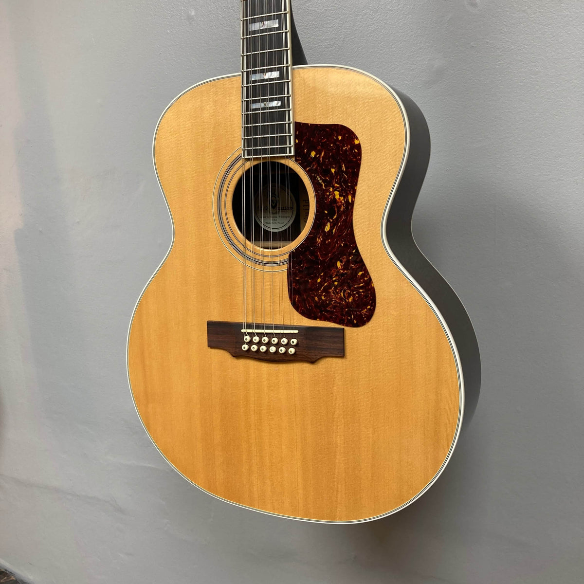 A Guild USA F-512 w/OHSC 2005 Circa 12-string jumbo guitar with a tortoiseshell pickguard hangs against a gray wall, showcasing classic Guild style.