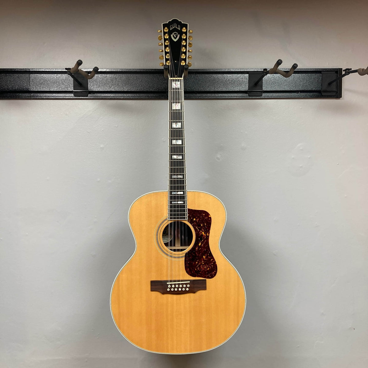 A Guild USA F-512 w/OHSC 2005 Circa 12-string acoustic guitar with a natural wood finish hangs on a black wall-mounted rack against a plain light-colored wall.
