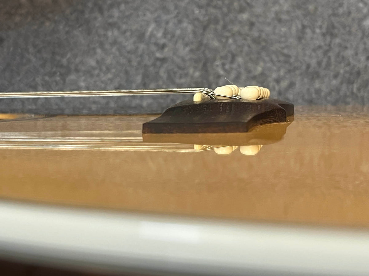Close-up of a Guild USA F-512 w/OHSC 2005 Circa 12-string jumbo acoustic guitar bridge with white pins and metal strings, by Guild, set against a blurred gray background. Crafted in Tacoma, Washington.