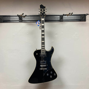 A Hagstrom Fantomen Custom Black w/case electric guitar, featuring a single cutaway, white binding, and dual Lundgren Design humbuckers, hangs on a wall-mounted guitar rack against a plain light-colored wall.