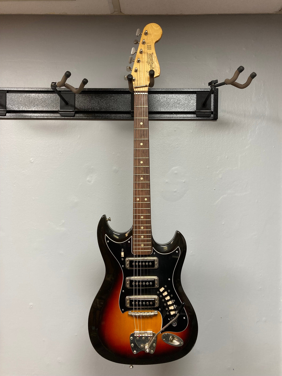 Hagstrom III 1960s Sunburst Electric Guitar with original case, displayed on a wall, showcasing vintage craftsmanship and minor scratches, available at Guitars on Main.