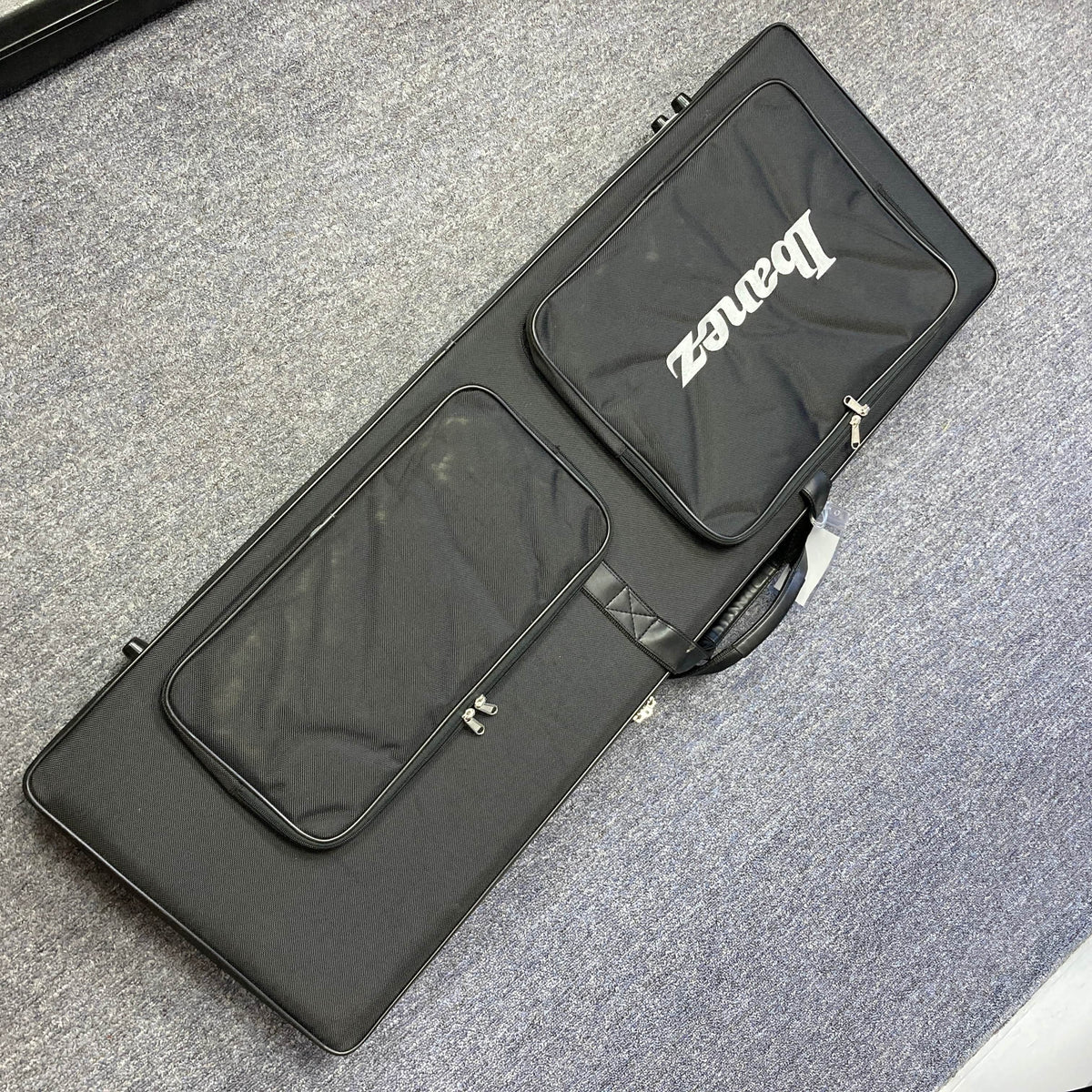 A black rectangular instrument case from Guitars on Main, ideal for the Ibanez JEM77P Steve Vai Signature electric guitar, sits on a gray carpeted floor and features two zippered pockets for convenience.