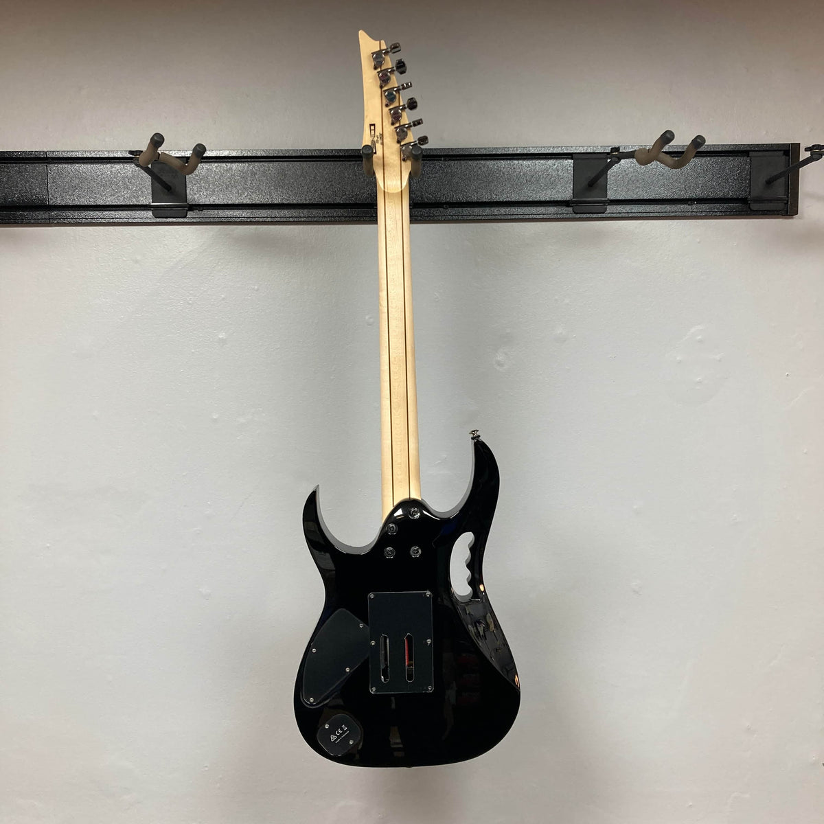 A black Ibanez JEM77P Steve Vai Signature by Guitars on Main hangs by its neck on a wall-mounted rack, viewed from the back against a plain light-colored wall.