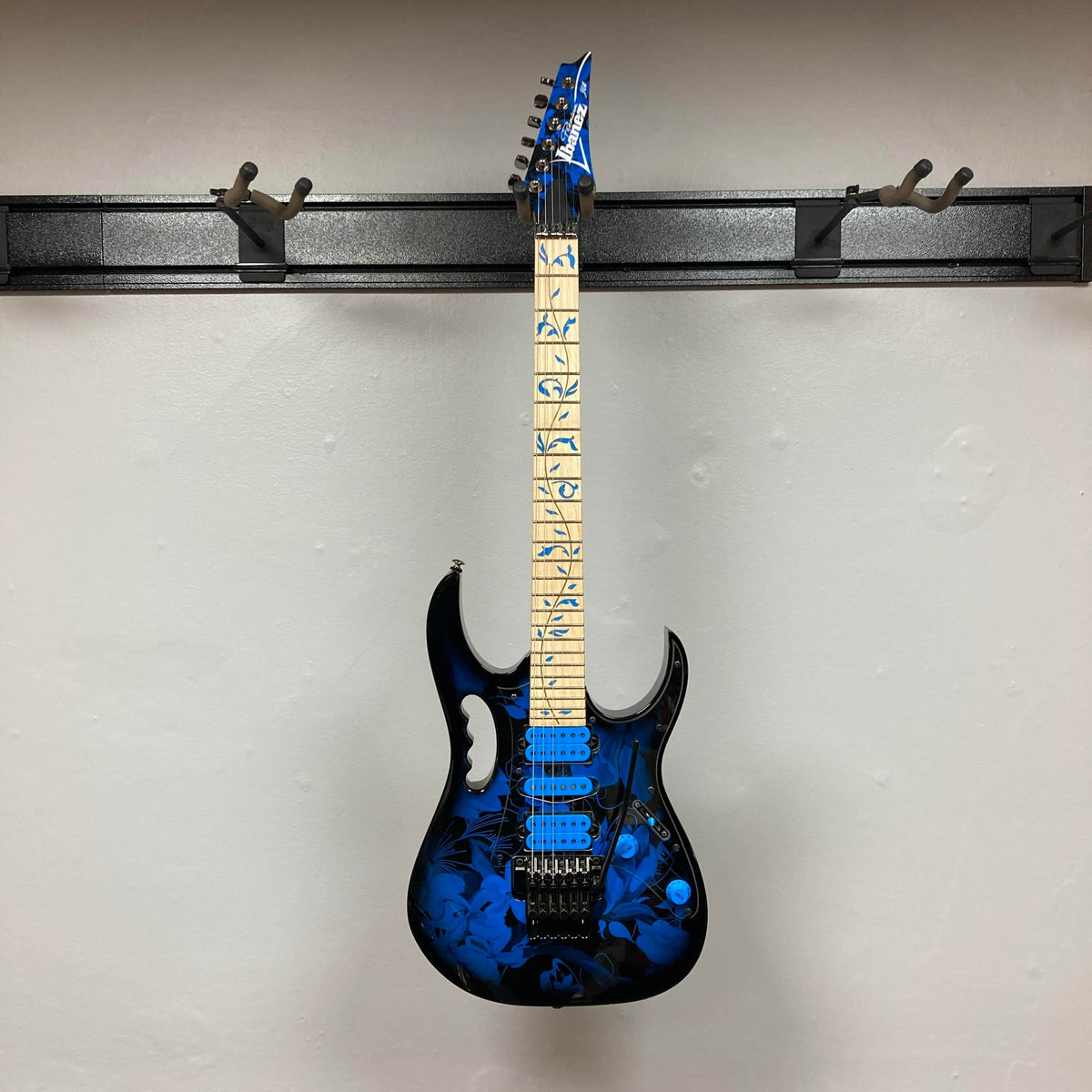 The Ibanez JEM77P Steve Vai Signature from Guitars on Main features a blue floral finish, maple neck with distinctive inlays, and matching blue headstock, displayed on a wall-mounted holder.