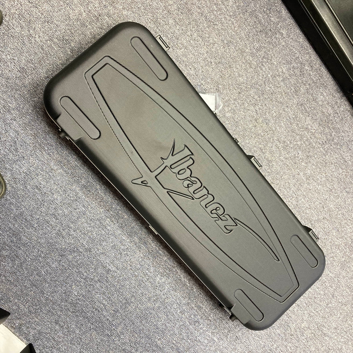 An Ibanez Prestige RG1570 Cosmic Blue guitar with a black hard case featuring the Ibanez logo, ideal for this Japanese-made model, is shown resting on a gray carpeted floor.