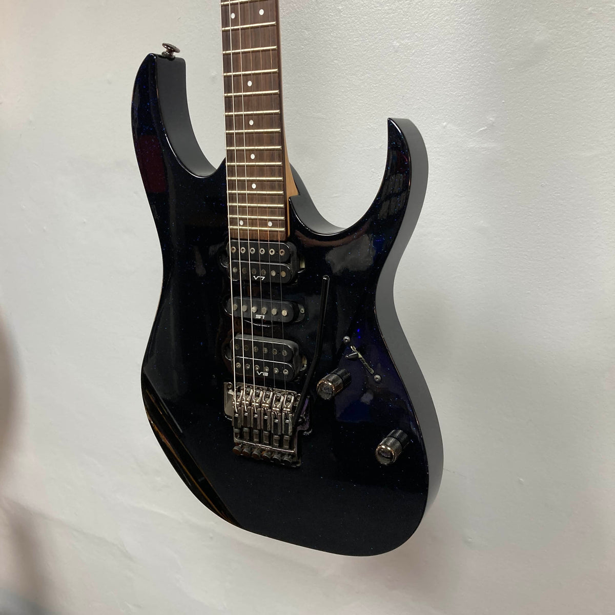 An Ibanez Prestige RG1570 Cosmic Blue electric guitar with a double-cutaway body and tremolo bridge hangs on a white wall.
