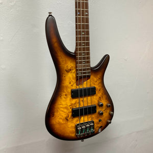 A close-up of the Ibanez SDGR SR500PB bass guitar by Ibanez, featuring a wood grain finish and multiple control knobs, displayed with its hard case against a plain white wall.