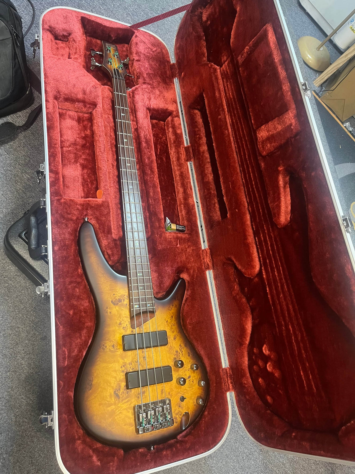 An Ibanez SDGR SR500PB bass guitar with flatwound strings rests in its red velvet-lined Ibanez hard case, open on a carpeted floor to showcase the instrument.