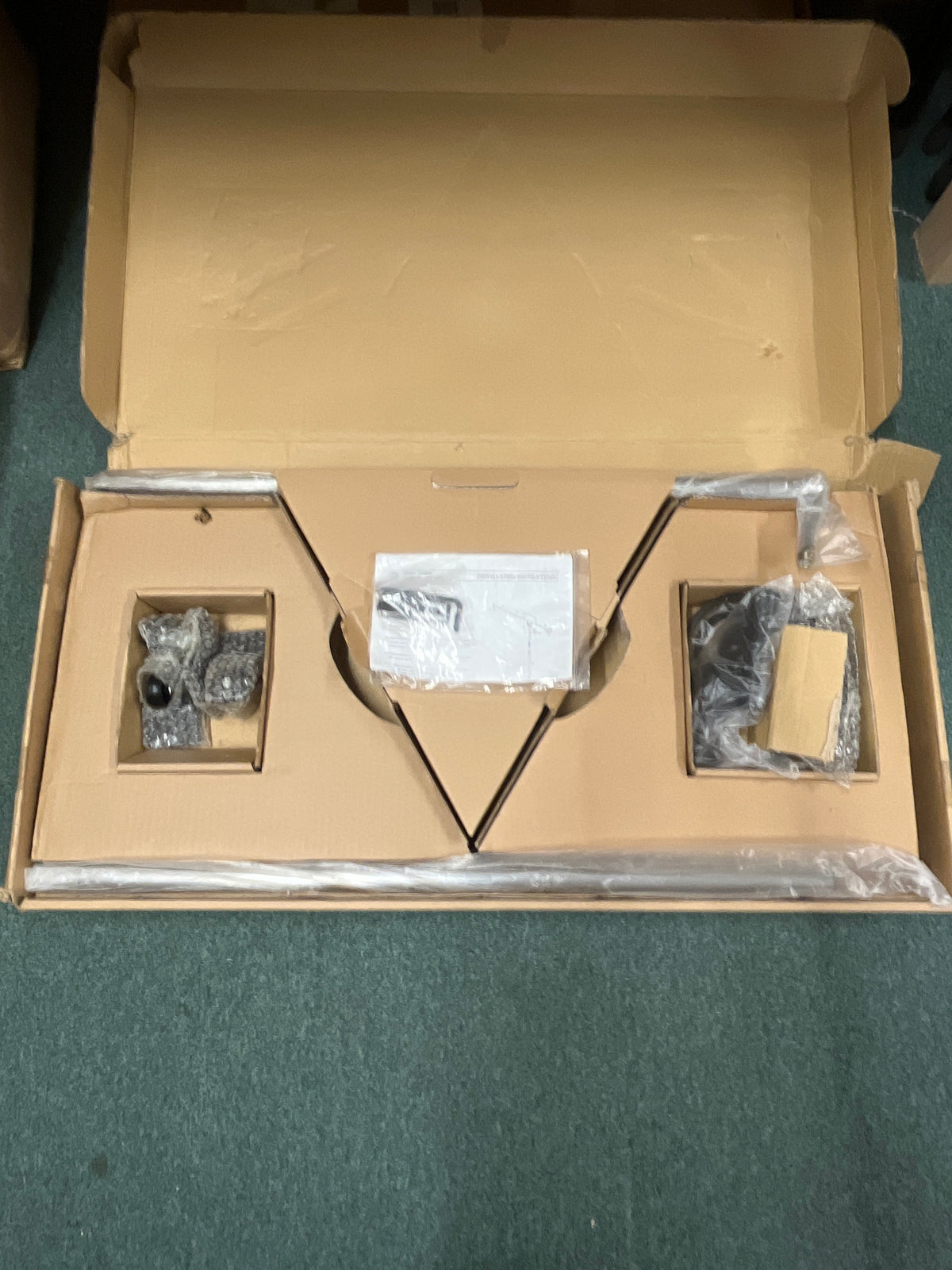 Opened cardboard box containing disassembled parts of an Atlas Sound SB36W Professional Studio Boom Microphone Stand, some wrapped in plastic and bubble wrap, placed on a green carpet.