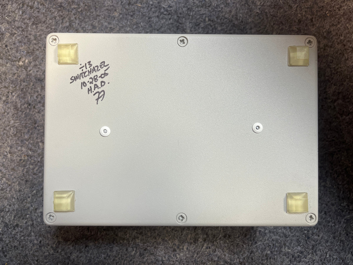 The Switchazel Boutique AB/Y Switcher with Tuner Out (Preowned) by Switchazel features a rectangular metal box, four rubber feet, and handwritten text on the bottom—perfect for guitar amp switching setups and eliminating ground loops.