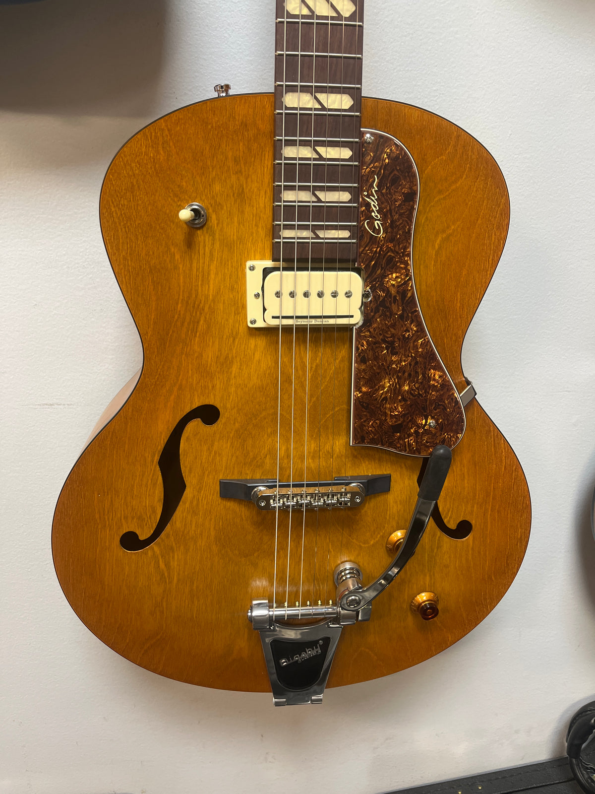 A Godin 5th Avenue Jumbo P-Rail Harvest Gold archtop guitar with a natural finish, single pickup, tortoiseshell pickguard, and Bigsby-style tailpiece hangs on a white wall.