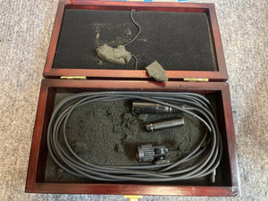 A wooden box with worn black foam holds a coiled audio cable and connectors, likely for the preowned Audix M1244-HC Miniature Condenser Hi-SPL Hypercardioid Microphone, resting on a textured gray surface.