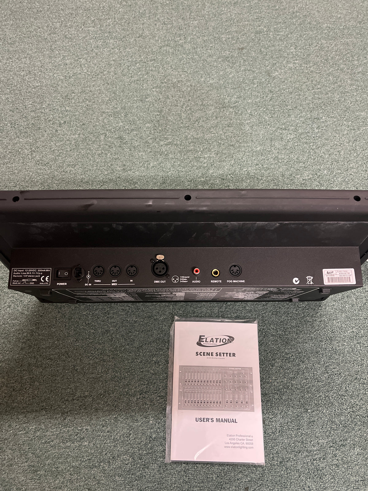 Back panel of the Elation Scene Setter 24-Channel DMX Dimmer Console by Elation, displaying multiple input/output ports, shown above its user manual on a carpeted floor—ideal for professional lighting control and scene programming.