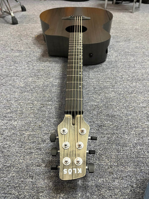 A Klos Grand Cutaway AE w/Fishman acoustic guitar lies flat on a carpeted floor, its carbon fiber body extending seamlessly from the headstock down the neck to the body.
