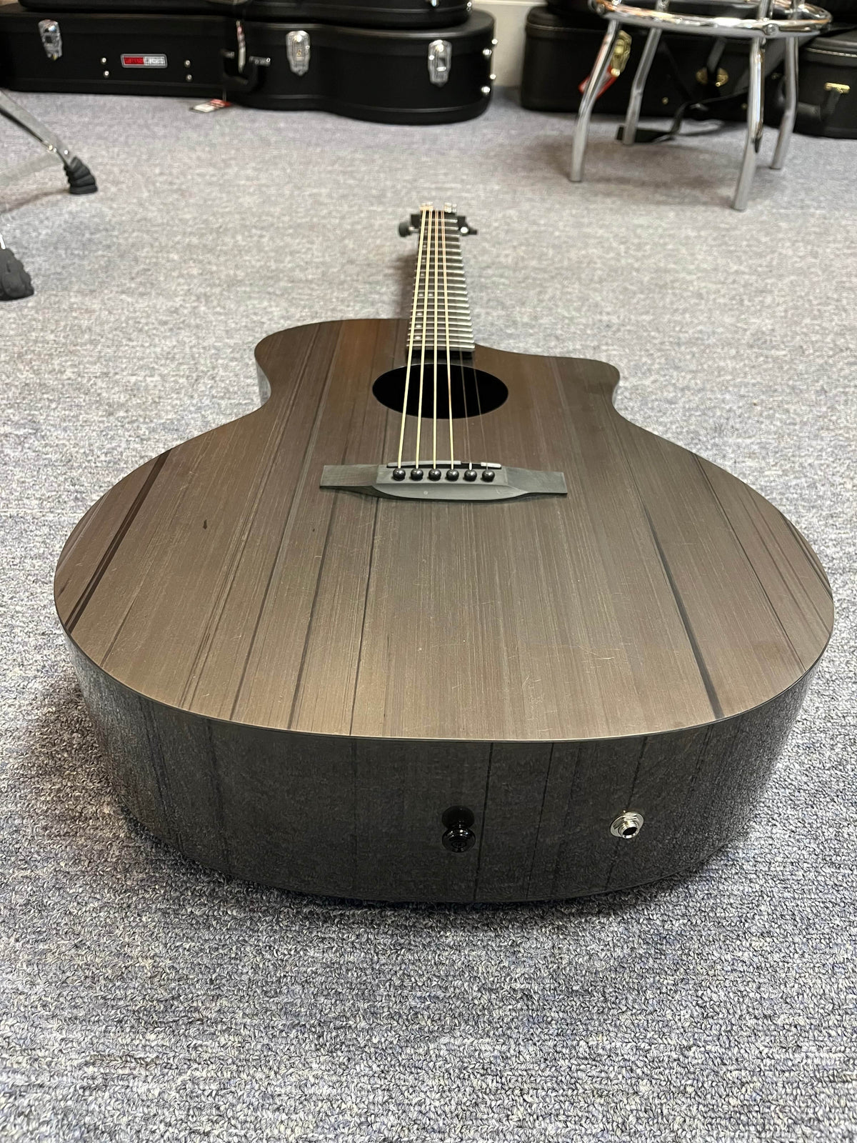 The Klos Grand Cutaway AE w/Fishman, a black acoustic guitar by Klos, rests on a gray carpeted floor with instrument cases and a stool in the background.