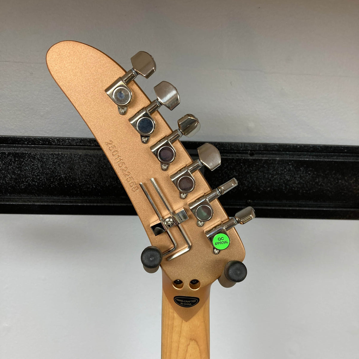 The back of a Kramer 84 HH Killswitch guitar headstock in Smoked Almond Metallic by Kramer shows six tuners, a barcode serial, “QC Passed” sticker, two Floyd Rose fine tuners, and a white wall with a black horizontal metal bar behind.