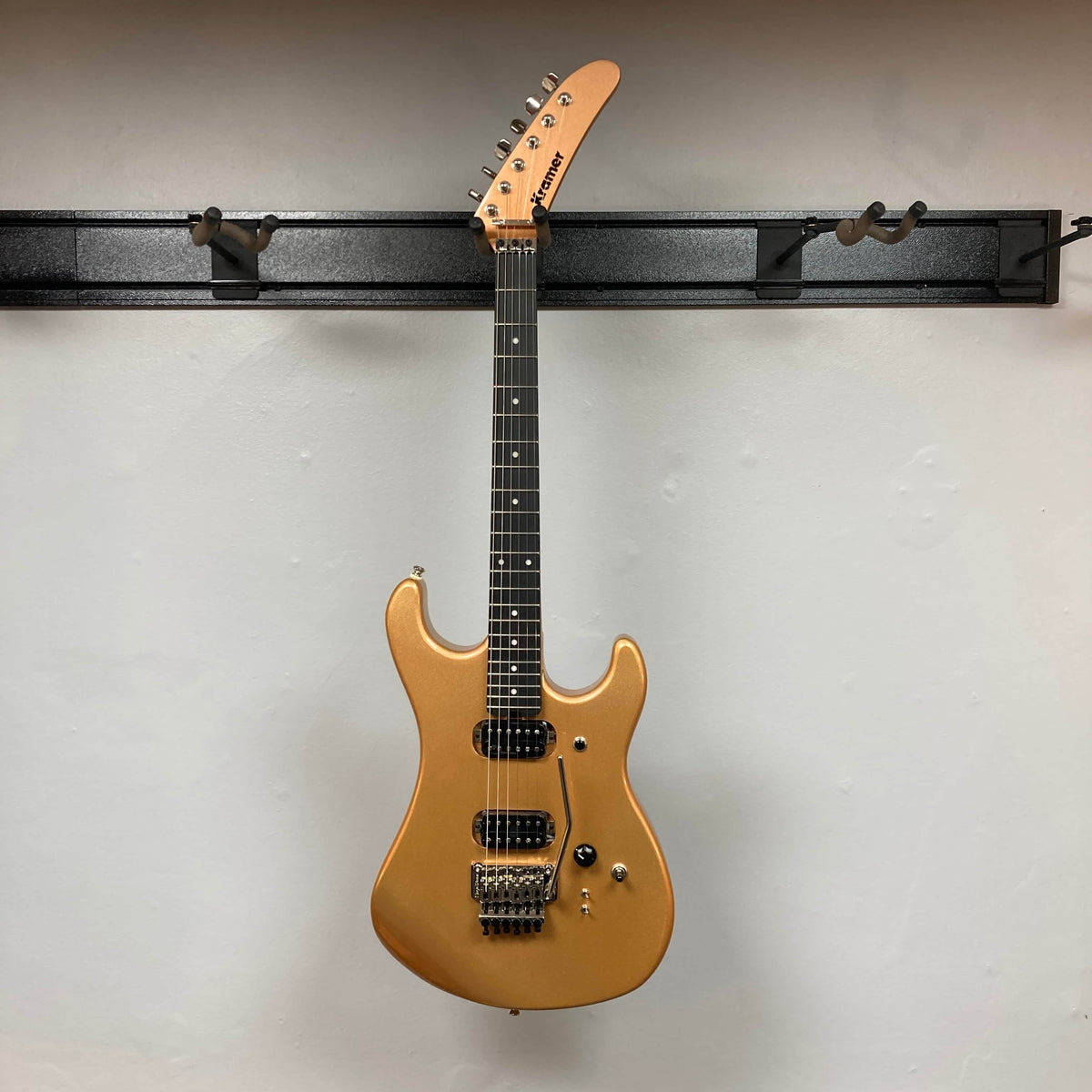 A Kramer 84 HH Killswitch electric guitar in Smoked Almond Metallic with a black fretboard is hanging on a black wall-mounted rack against a plain white wall.