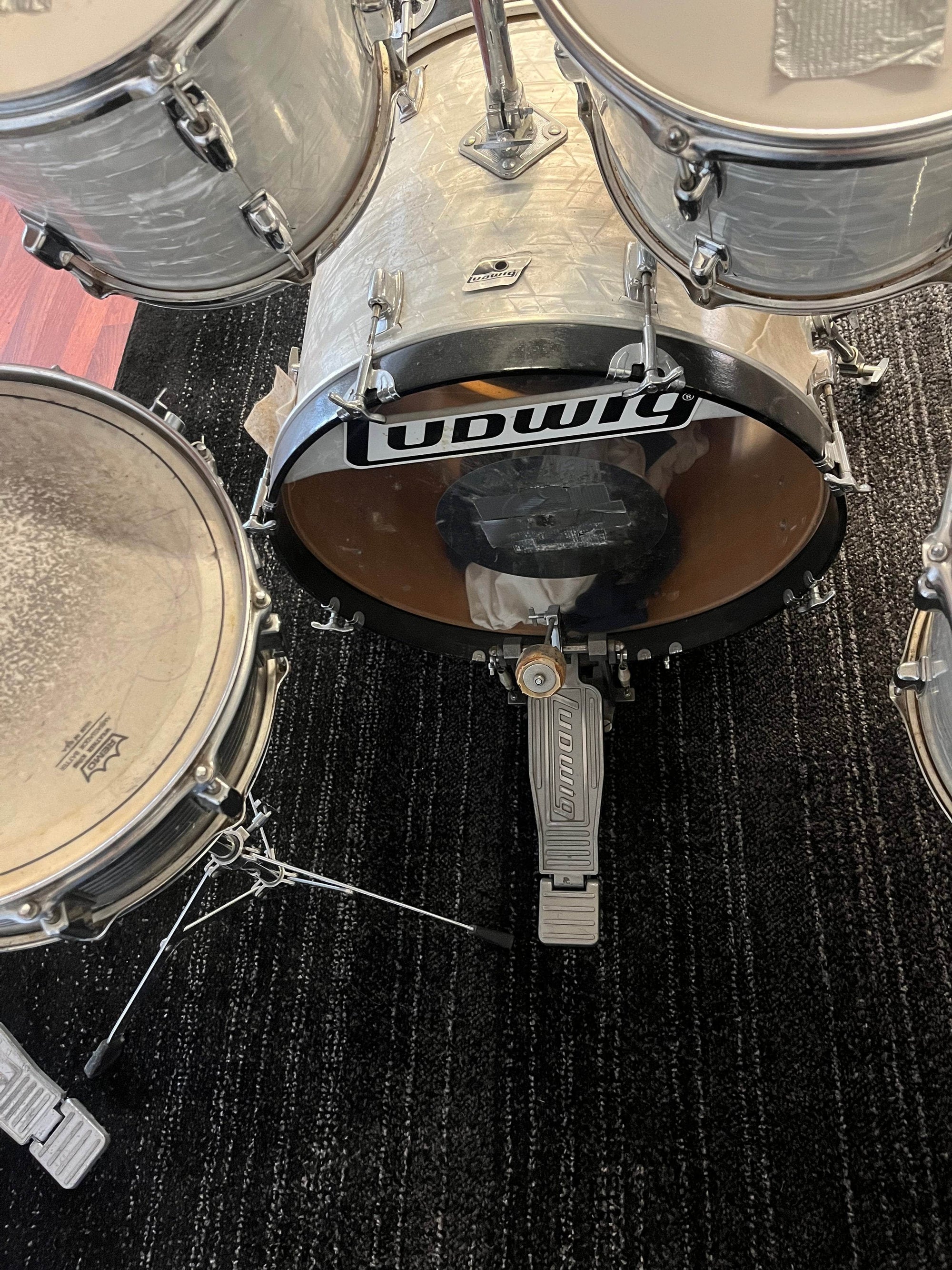 A vintage Ludwig Classic 1980s Drum Kit in White Marine Pearl is set on a dark carpet before a plain gray partition, cymbals included. A yellow flower rests atop the bass drum, adding charm to this classic Ludwig set.
