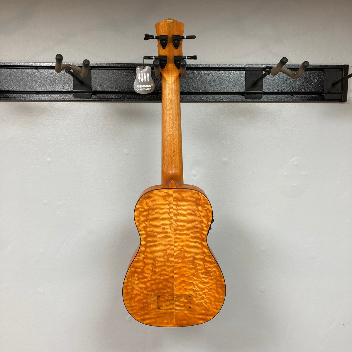 Luna Bari-Bass Quilt Ash Top Bass Ukulele w/Preamp, featuring a quilt ash body, mahogany neck, walnut fingerboard, and open gear tuners, displayed on a wall.