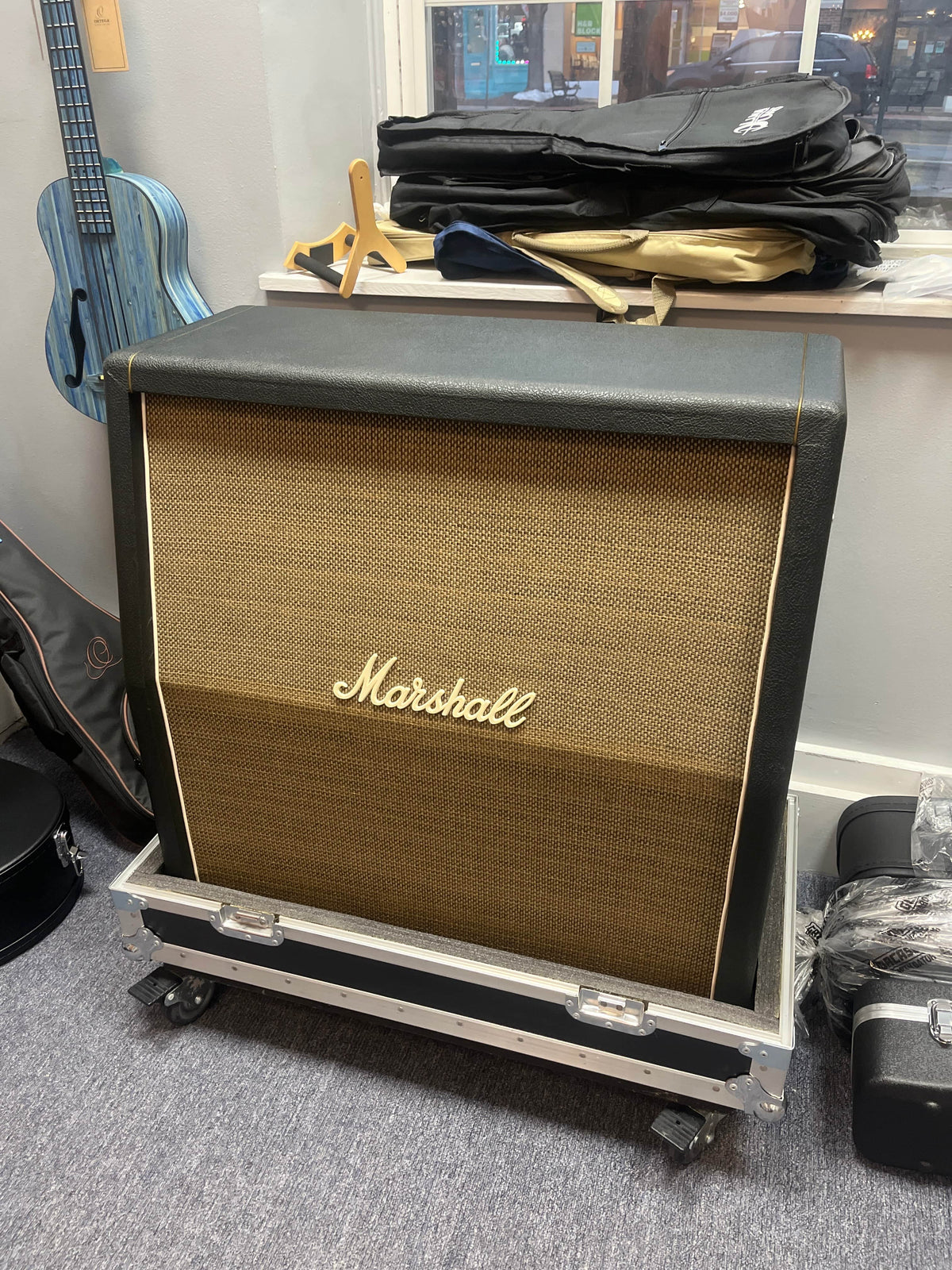 A 1969 Marshall 4x12 Cabinet with matching Celestion T1281 Greenbacks sits on a rolling case in a room filled with instrument cases and a blue guitar hanging on the wall.