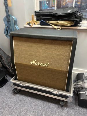 A 1969 Marshall 4x12 Cabinet with matching Celestion T1281 Greenbacks sits on a rolling case in a room filled with instrument cases and a blue guitar hanging on the wall.