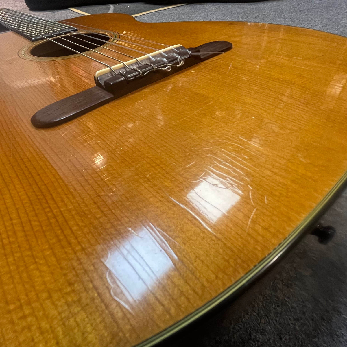 Close-up of a Martin 00-18G Classical Guitar (c. 1961–1962) showing light scratches and finish cracks on the wood surface, with the bridge and strings visible.