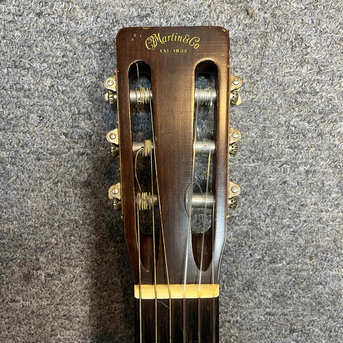 Close-up of the headstock of a Martin 00-18G Classical Guitar, c. 1961–1962 by Martin, featuring six tuning pegs and nylon strings set against a textured gray background.