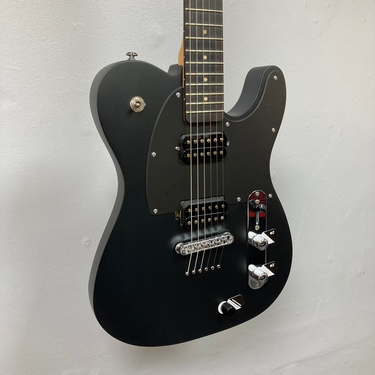 Michael Kelly Hybrid 55T Black Satin Blem electric guitar with dual humbucker pickups and Fishman Powerbridge, mounted on a white wall.