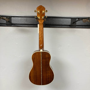 The Oscar Schmidt OU280SWK Concert Ukulele, by Oscar Schmidt, hangs on a wall mount. Its back has a glossy finish, central inlay strip, abalone binding, and gold tuning pegs. Comes with case; used consign.