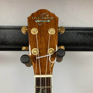 Close-up of the Oscar Schmidt OU280SWK Concert Ukulele headstock with four tuning pegs, gold hardware, abalone binding, visible strings, and the Oscar Schmidt logo. Shown mounted on a black wall hanger. Case included; used consign.