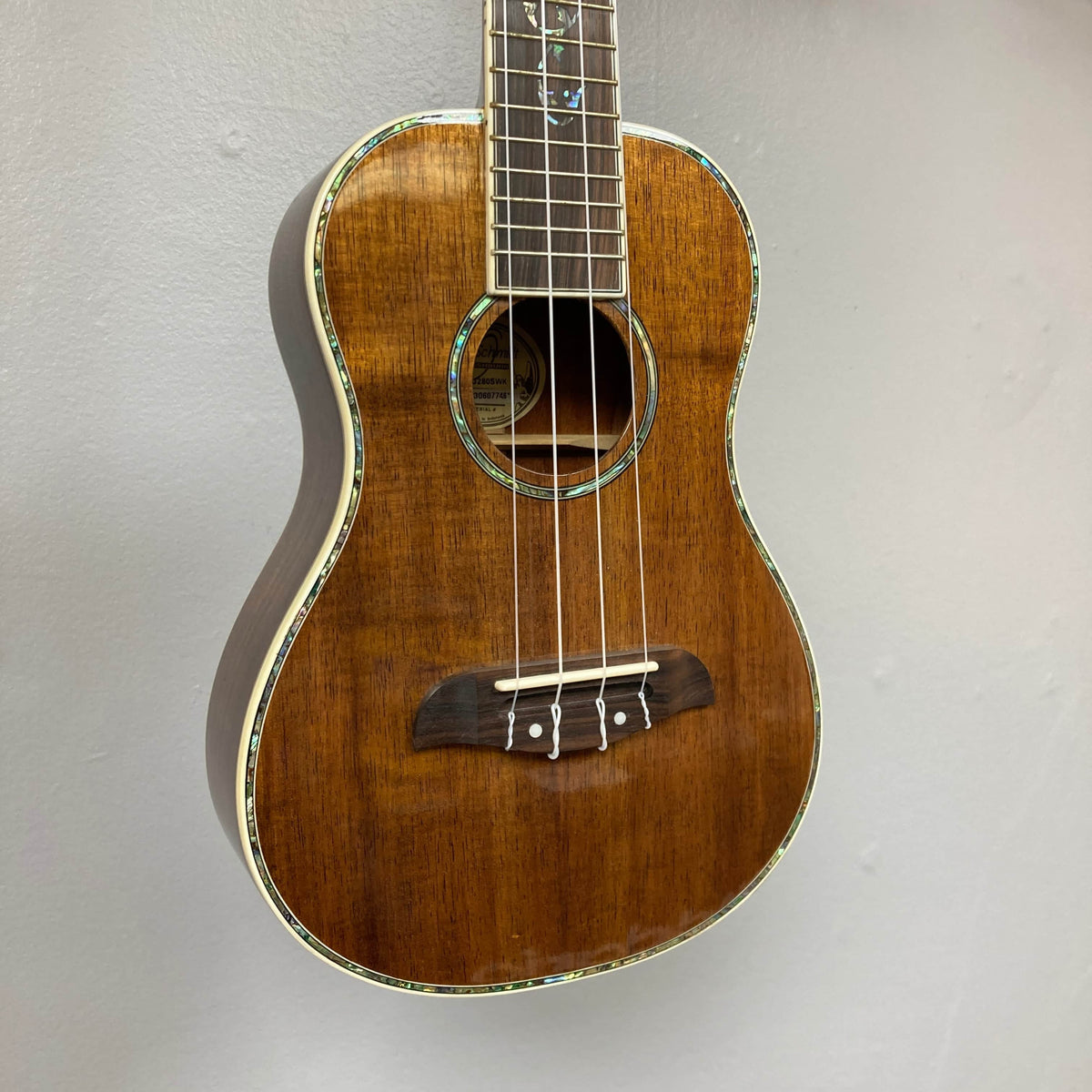 The Oscar Schmidt OU280SWK Concert Ukulele, a brown solid koa model with four strings and abalone binding, features a glossy finish and hangs on a light gray wall.